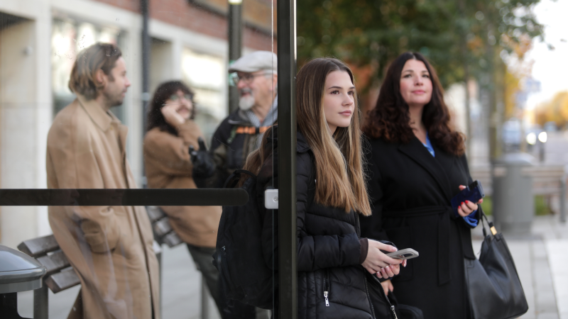 Fem personer står och väntar på bussen vid en busshållsplats.