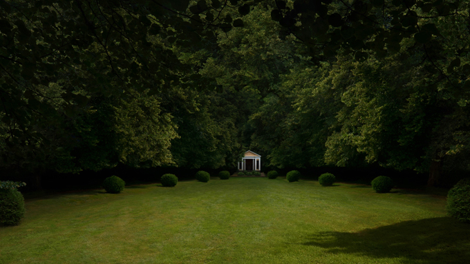 Konstnären Lovisa Ringborgs verk Portal. Öppen gräsyta omringad av lövskog. I mitten av bilden, vid skogskanten, skymtar en liten byggnad.