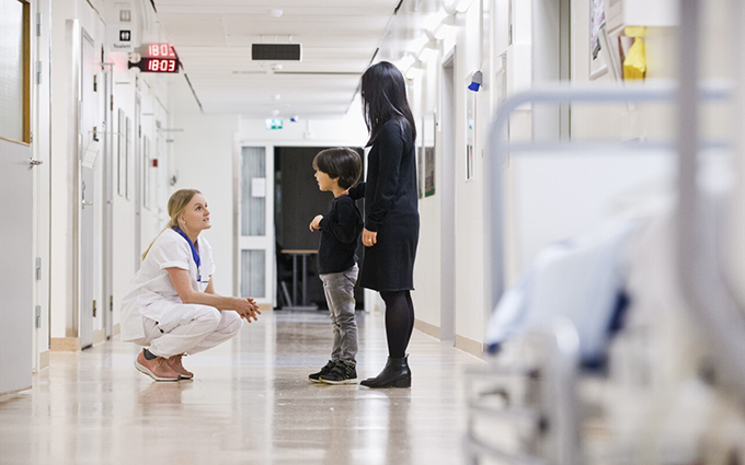 Vårdpersonal på huk framför barn och förälder i korridor.
