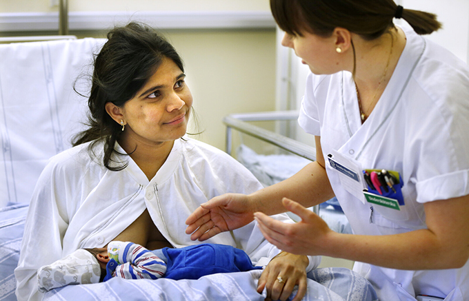 Mamma, ammande spädbarn och vårdpersonal vid sängen.