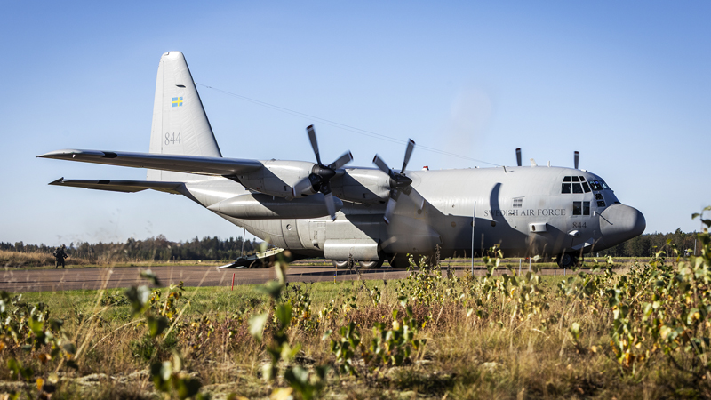 Ett stort grått transportflygplan på en landningsbana. I förgrunden syns gröna växter.