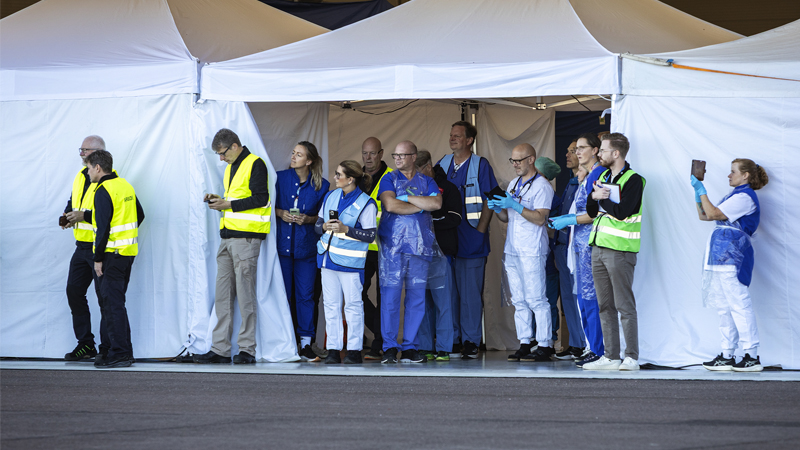 Ett vitt tält med en öppning. Utanför står ett stort antal personer och tittar åt samma håll. Flera av dem är klädda i hälso- och sjukvårdens blåa och vita kläder.