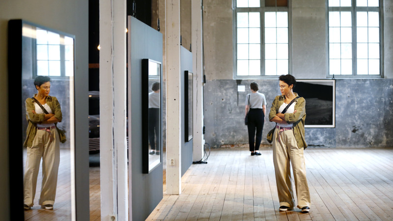 Konstutställning. Två kvinnor, en i förgrunden och en längre bort tittar på stora tavlor.