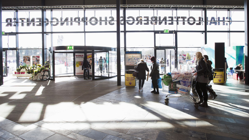 Entré i glasparti där skylten Charlottenbergs köpcentrum syns genom glaset bakifrån. Människor med kundvagnar är på väg ut genom entrésnurran.