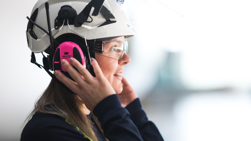 Profil av kvinna med  vit hjälm, skyddsglasögon och rosa hörselkåpor som håller händerna på hörselkåporna.
