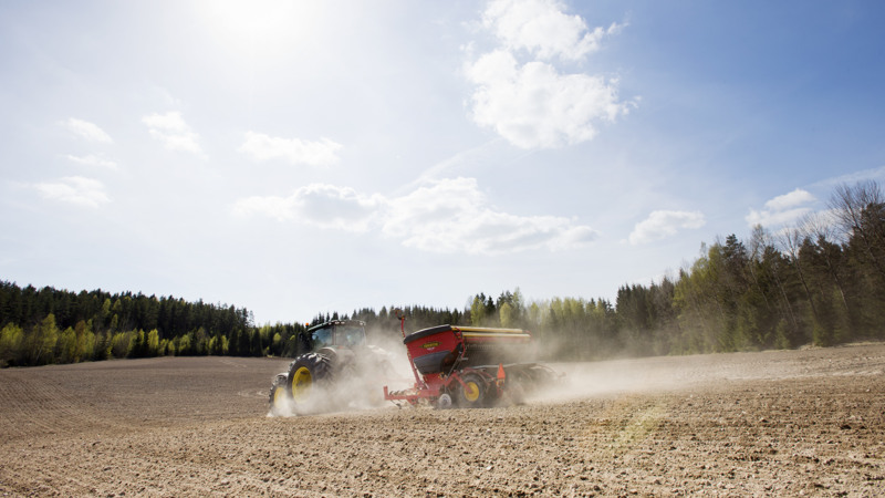 Traktor med såmaskin kör på åker med ett dammoln efter en solig dag. 