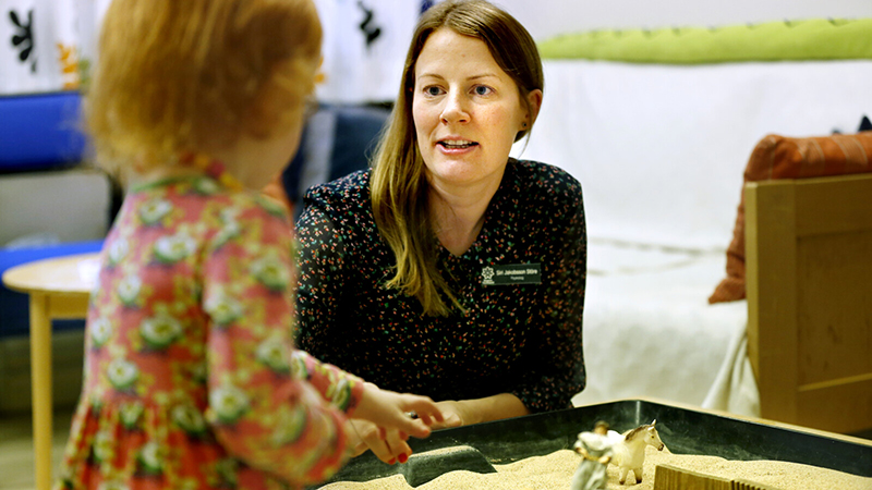 Kvinna pratar med ett barn vid en inomhussandlåda.