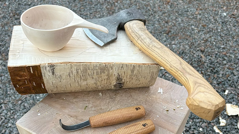 En kåsa i trä tillsammans med verktygen yxa och kniv.