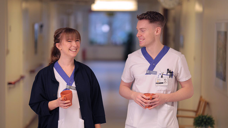 Två dietister i vita arbetskläder med varsin kopp i handen i en korridor.