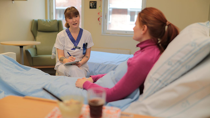 Kvinna i arbetskläder sitter och antecknar bredvid sängliggande patient.