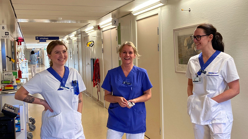 Tre glada kvinnor i arbetskläder i sjukhuskorridor.