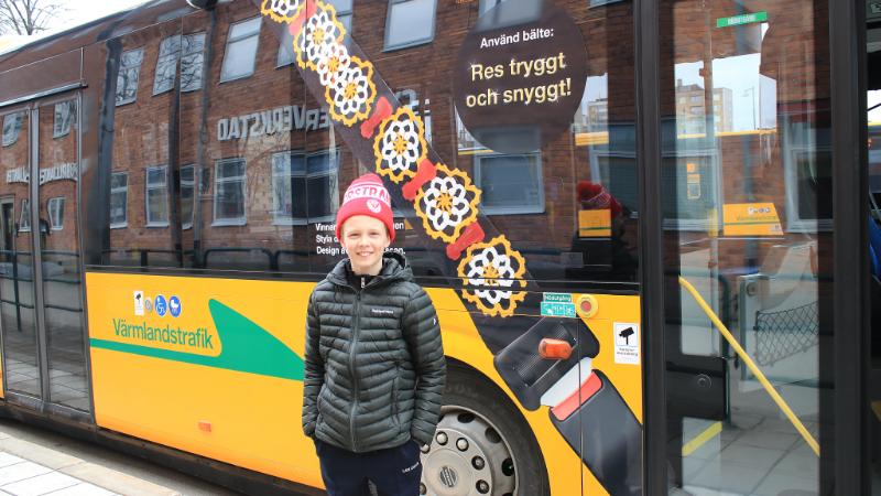 Algot Eriksson framför en gul Värmlandstrafiksbuss där en stor klisterdekal med hans vinnarbussbälte finns uppsatt.