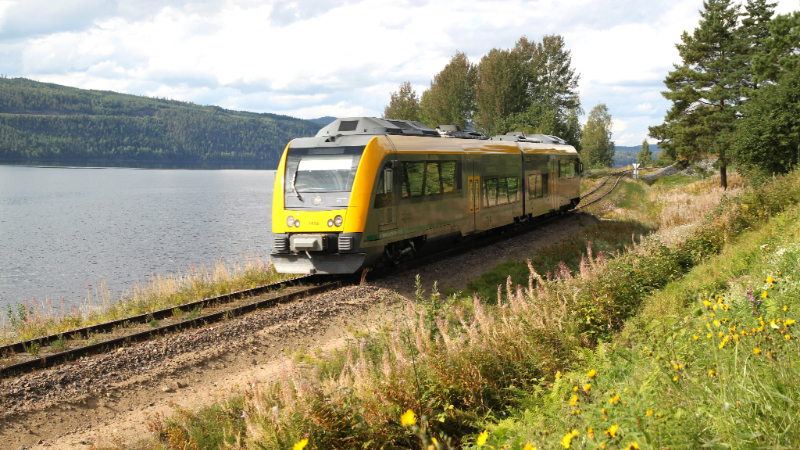 Ett gult och silvrigt Itino-tåg kör fram på Fryksdalsbanan i vacker natur. Tåget är omgivet av gräs och vatten.