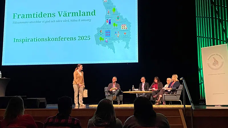 En person står på en scen och pratar till fem personer som sitter på scenen och samtalar.