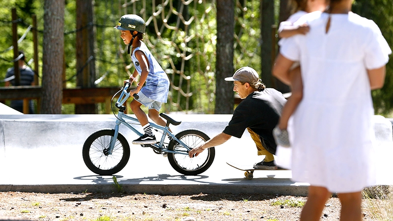 Barn som cyklar och drar sin pappa på cykel bakom sig. 
