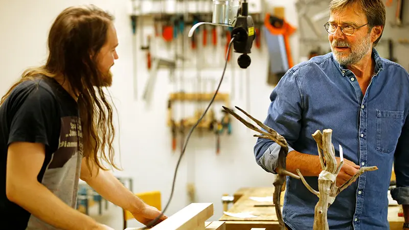 Två personer står i en verkstad med träfigurer framför sig. 