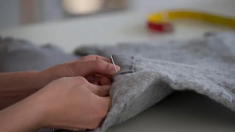 Närbild på händer som syr i grått ylletyg. 