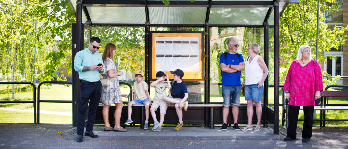 En man, en mamma med två pojkar, ett äldre par och en kvinna står och väntar vid en busshållsplats. 