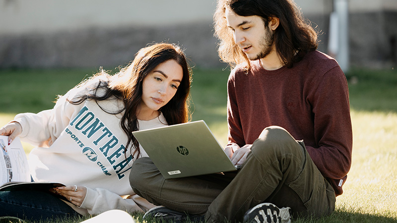 Två personer sitter utomhus och tittar på en laptop som ligger i den ena personens knä.