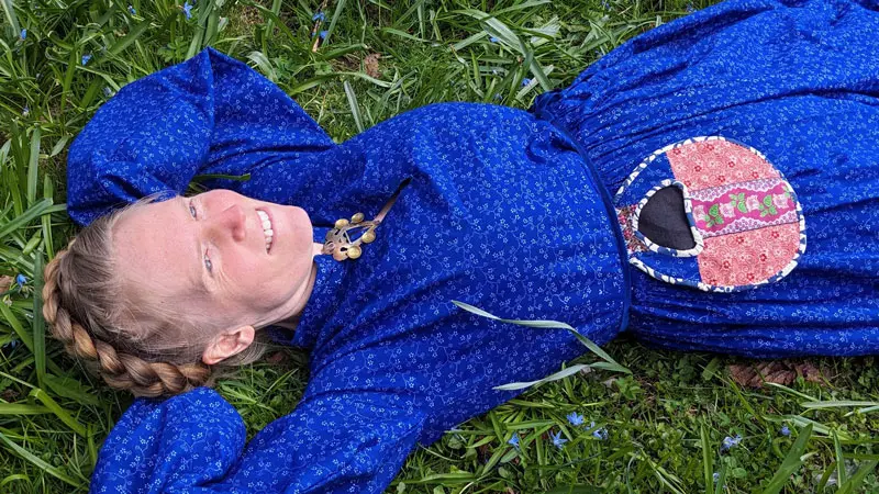 Kvinna i blå folkdräkt ligger med händerna bakom huvudet på en gräsmatta.