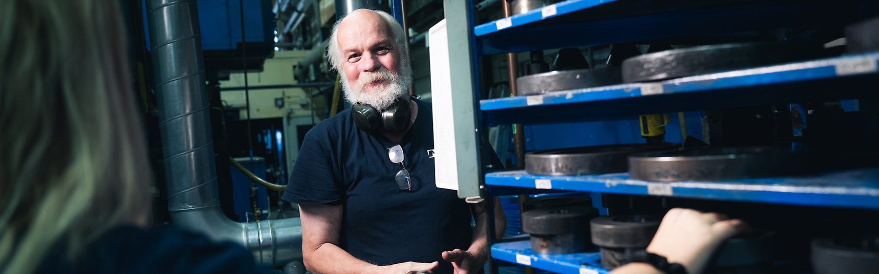 Glad man med grått hår och grått skägg, med hörselkåpor runt halsen, vänd mot fotograf i en verkstadsmiljö.